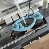 Blue fish-shaped plating yarn holder mounted on a Brother double bed knitting machine